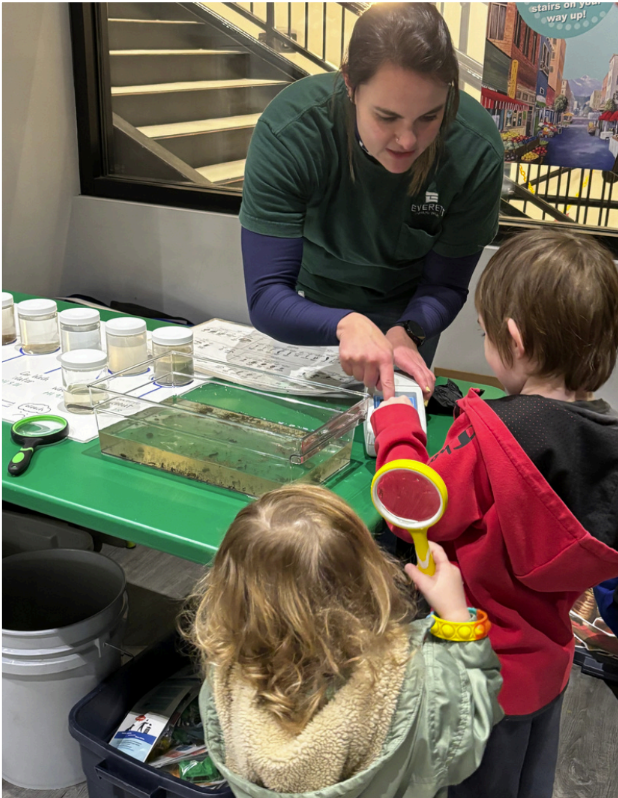 An educator demonstrates stream science at a table while two children examine water samples with magnifying tools.